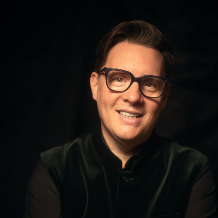 A smiling person with short brown hair, wearing glasses and a dark, buttoned-up velvet jacket, sits against a dark, softly lit background.