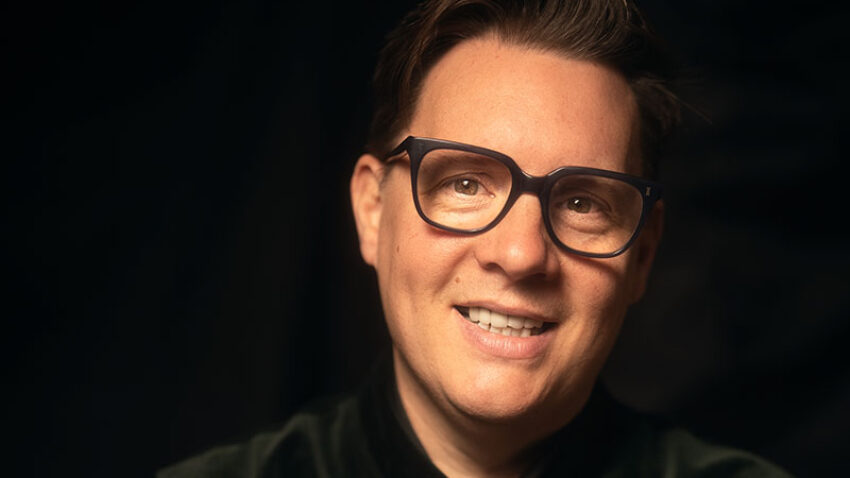 A smiling person with short brown hair, wearing glasses and a dark, buttoned-up velvet jacket, sits against a dark, softly lit background.