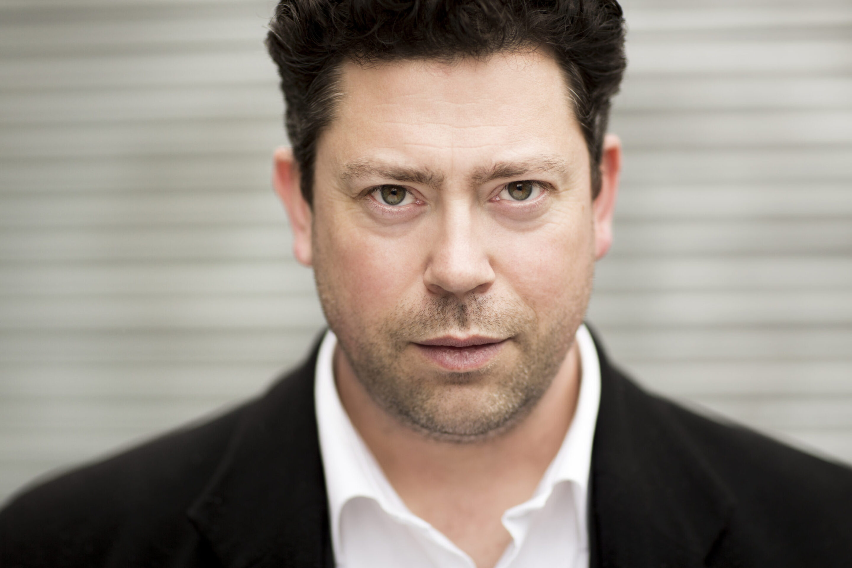 A man with short, dark curly hair wearing a black blazer and white shirt stares directly at the camera with a serious expression. The background is blurred grey horizontal lines.