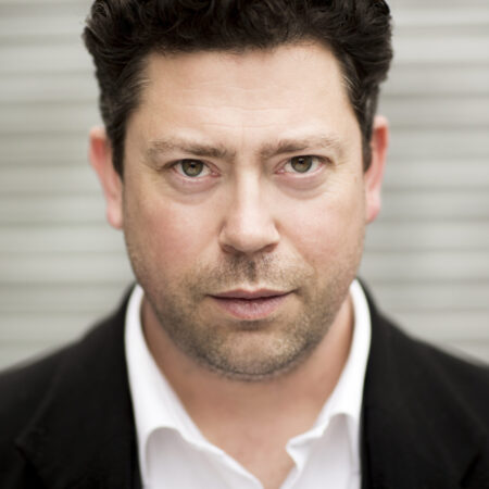A man with short, dark curly hair wearing a black blazer and white shirt stares directly at the camera with a serious expression. The background is blurred grey horizontal lines.