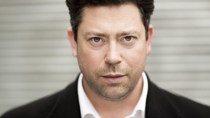 A man with short, dark curly hair wearing a black blazer and white shirt stares directly at the camera with a serious expression. The background is blurred grey horizontal lines.