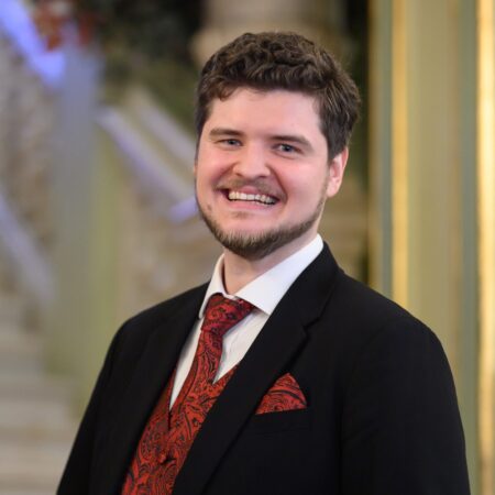 Egor Zhuravskii, a smiling man with short brown hair and a trimmed beard, wears a black suit, white shirt, and red patterned tie with a matching pocket square as he stands indoors before an elegant staircase.