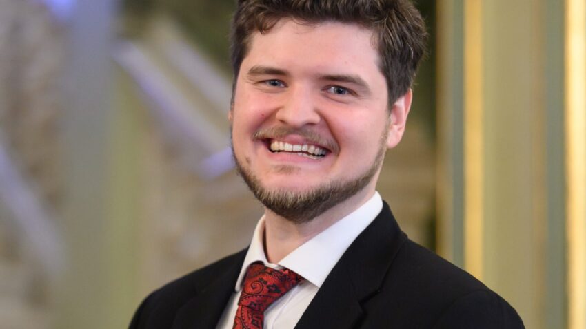 Egor Zhuravskii, a smiling man with short brown hair and a trimmed beard, wears a black suit, white shirt, and red patterned tie with a matching pocket square as he stands indoors before an elegant staircase.