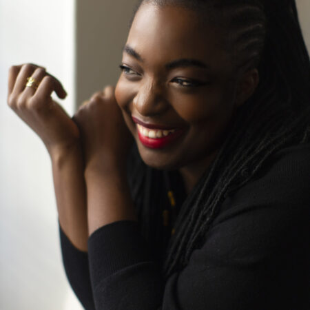 A woman with long plaited hair and bright red lipstick is smiling and looking to the side, resting her arms on a surface. She is wearing a black top and appears happy and relaxed.