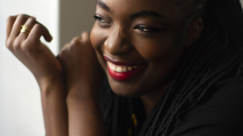 A woman with long plaited hair and bright red lipstick is smiling and looking to the side, resting her arms on a surface. She is wearing a black top and appears happy and relaxed.
