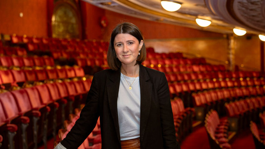 Helen Shute, stands smiling in an ornate, empty London Coliseum theatre with shoulder-length brown hair, wearing a black blazer and light top amidst rows of red seats and decorative lighting overhead.