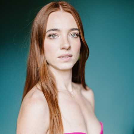Joanne Evans, with long auburn hair, wears a strapless bright pink dress and looks slightly past the camera. The background features a smooth gradient of blue and green tones.