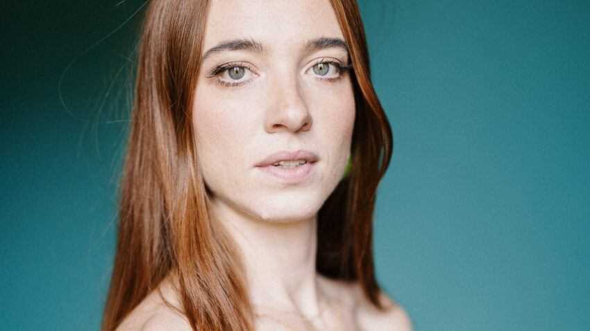 Joanne Evans, with long auburn hair, wears a strapless bright pink dress and looks slightly past the camera. The background features a smooth gradient of blue and green tones.