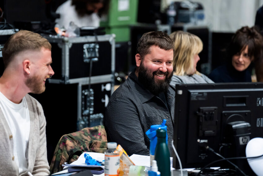 Two men sit at a desk with monitors and papers, smiling and talking. Behind them are electronic equipment cases and other people, suggesting a busy work or production environment.