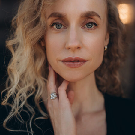 Kristina Stanek, with wavy blonde hair and blue eyes, gazes at the camera. She wears subtle make-up, a black top, and a diamond ring on her finger, which gently touches her neck against a softly blurred background.
