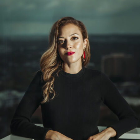 A woman, Lauren Snouffer, with wavy light brown hair and red earrings stands on a balcony in a black long-sleeved dress, looking at the camera. A blurred cityscape and cloudy sky serve as the background.