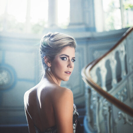 Lauren Fagan, in a strapless, elegant dress, stands on a grand staircase with ornate railings, looking over her shoulder as sunlight streams through large windows, softly illuminating her and the intricate surroundings.