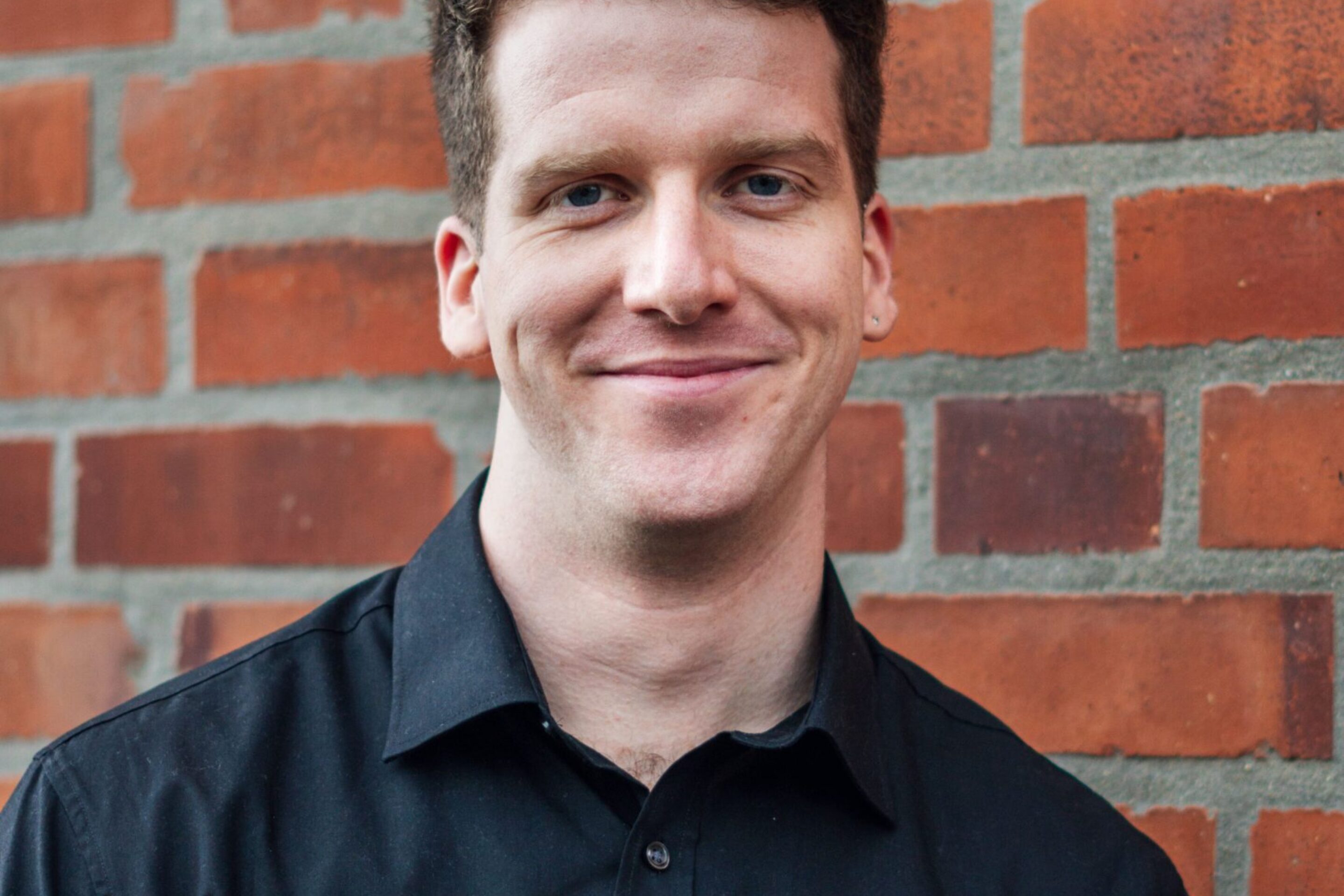 Liam James Karai, a man with short curly brown hair wearing a black button-up shirt, is standing and smiling in front of a red brick wall.