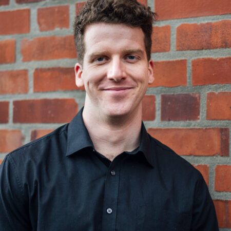 Liam James Karai, a man with short curly brown hair wearing a black button-up shirt, is standing and smiling in front of a red brick wall.