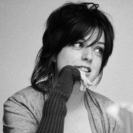A young woman with dark, tousled hair and a fringe looks to the side, lightly biting her finger. She wears a long-sleeved shirt and a cardigan, appearing thoughtful or pensive. The photo is in black and white.