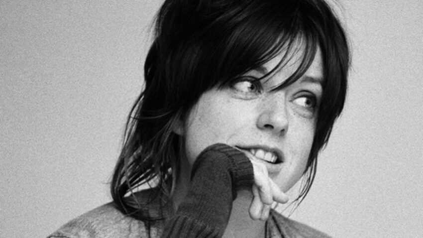 A young woman with dark, tousled hair and a fringe looks to the side, lightly biting her finger. She wears a long-sleeved shirt and a cardigan, appearing thoughtful or pensive. The photo is in black and white.