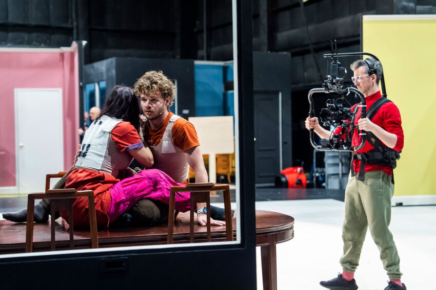 A camera operator films two actors in costume sitting closely at a table on a brightly lit set with colourful walls, capturing an intense moment in their performance.