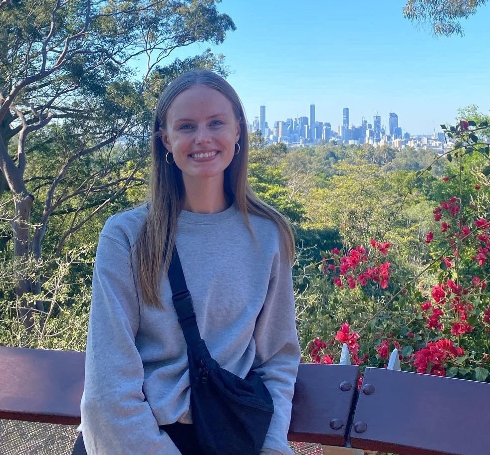 A smiling woman, Jess, with long straight hair, wearing a grey sweatshirt and black crossbody bag, sits on a bench outdoors. Behind her are trees, red flowers, and a distant city skyline under a clear blue sky—perfect for a chat in Issue 15.