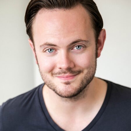 Nick Pritchard, a man with light skin, short brown hair, and a trimmed beard, is smiling slightly. He is wearing a dark V-neck jumper and faces the camera against a neutral, light background.