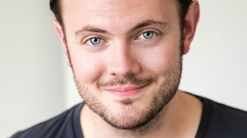 Nick Pritchard, a man with light skin, short brown hair, and a trimmed beard, is smiling slightly. He is wearing a dark V-neck jumper and faces the camera against a neutral, light background.