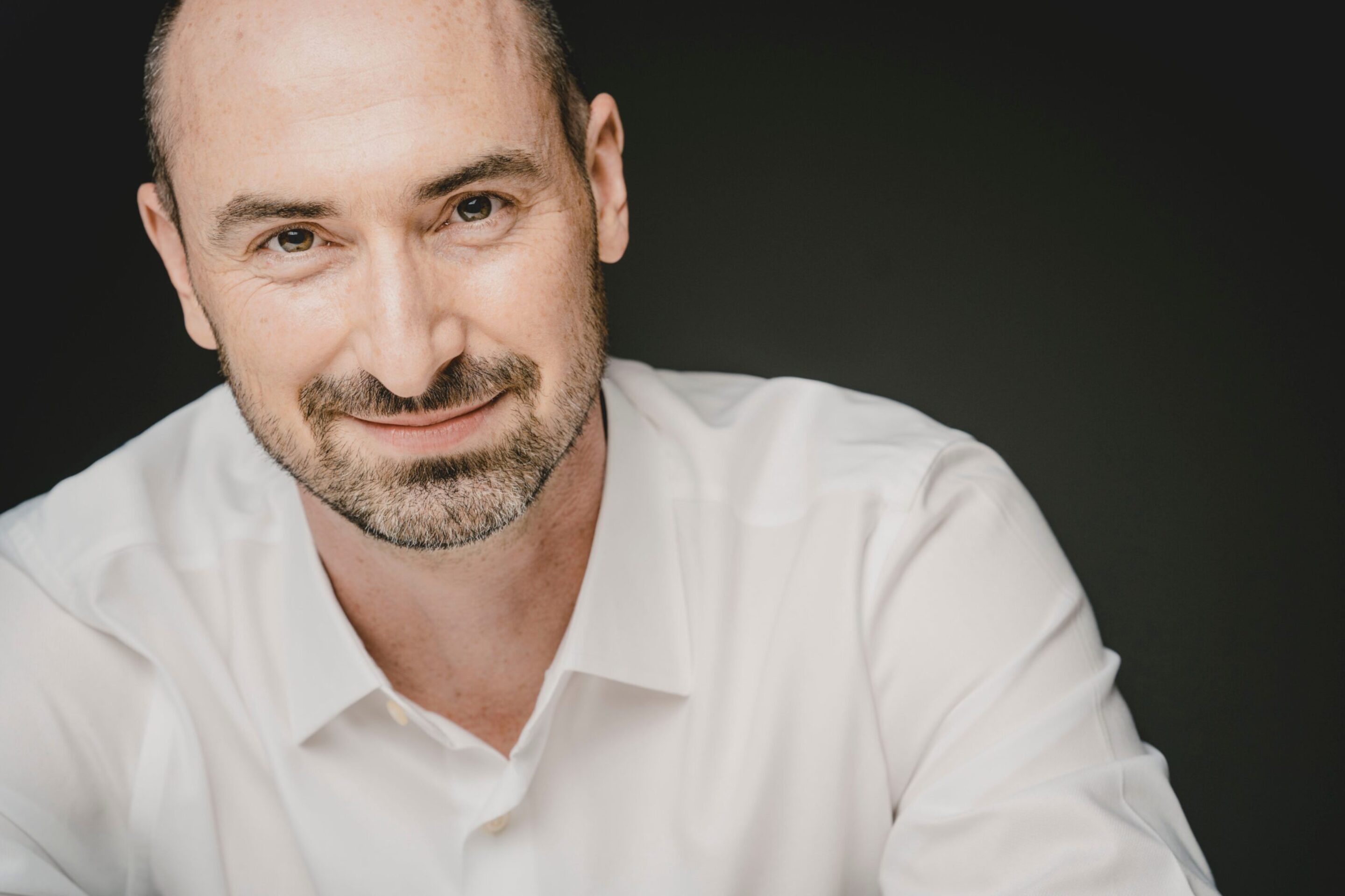 Peter Whelan, a smiling man with a shaved head and beard, wearing a white dress shirt, poses against a dark background.
