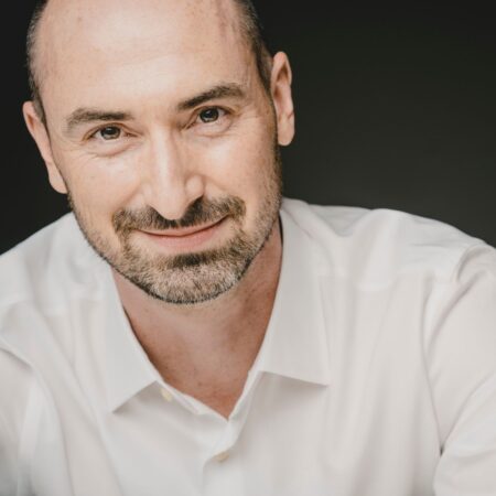 Peter Whelan, a smiling man with a shaved head and beard, wearing a white dress shirt, poses against a dark background.
