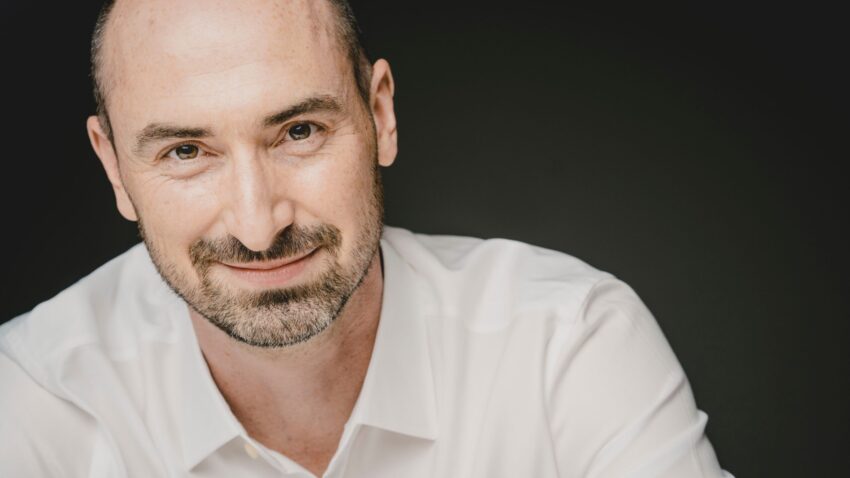 Peter Whelan, a smiling man with a shaved head and beard, wearing a white dress shirt, poses against a dark background.