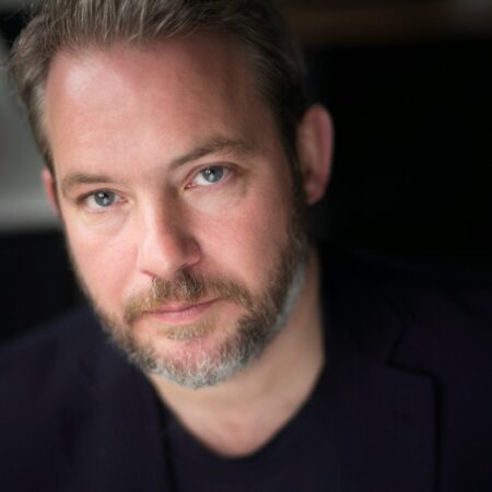 A man with light skin, short brown hair, and a neatly trimmed grey beard looks into the camera with a neutral expression. He is wearing a black shirt and a dark jacket, and the background is softly blurred.