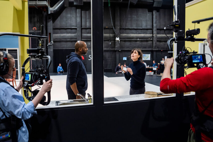 Two people are filmed by a crew inside a studio, standing at a counter and talking, while camera operators capture the scene from both sides through large glass windows. The set has industrial and theatrical elements.