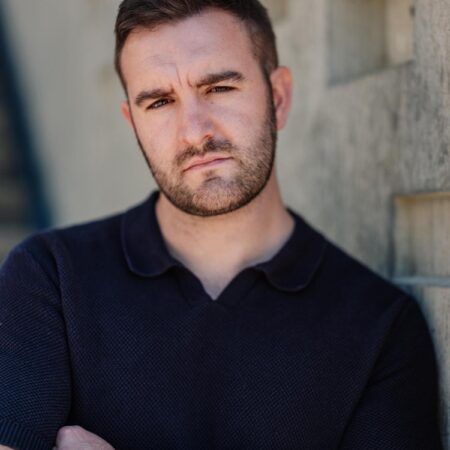 Steffan Lloyd Owen, a man with short brown hair and a trimmed beard, wearing a dark collared shirt, stands with arms folded against a concrete wall with square cut-outs, looking seriously at the camera.