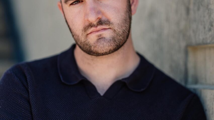Steffan Lloyd Owen, a man with short brown hair and a trimmed beard, wearing a dark collared shirt, stands with arms folded against a concrete wall with square cut-outs, looking seriously at the camera.
