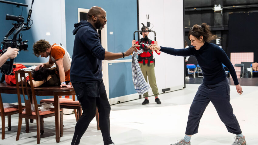 Two people in casual clothes rehearse a fight scene on a set, holding prop weapons and facing each other. In the background, others watch, including a camera operator filming the action. The scene appears staged indoors.