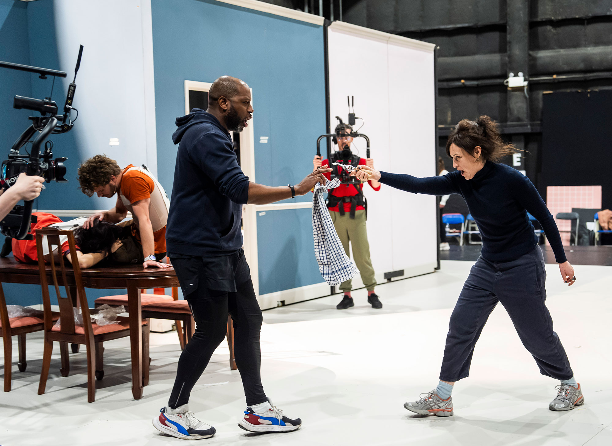 Two people in casual clothes rehearse a fight scene on a set, holding prop weapons and facing each other. In the background, others watch, including a camera operator filming the action. The scene appears staged indoors.