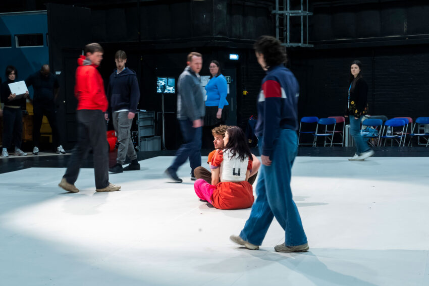 A group of people walk across a large indoor space with white flooring, whilst two people sit on the floor in the centre. Blue chairs are lined up against the back wall. The atmosphere appears casual and informal.