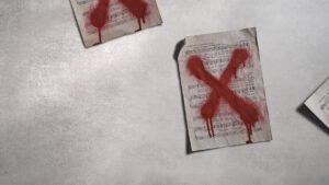 Three sheets of music lie on a grey surface, each marked with a large, red, painted X that appears to be dripping, suggesting rejection or censorship.