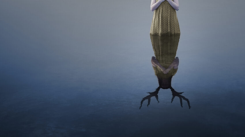 A person in a green dress stands by still water, hands clasped. Their reflection reveals antlers in place of a human head, evoking the haunting duality found in Iphigénie en Tauride.