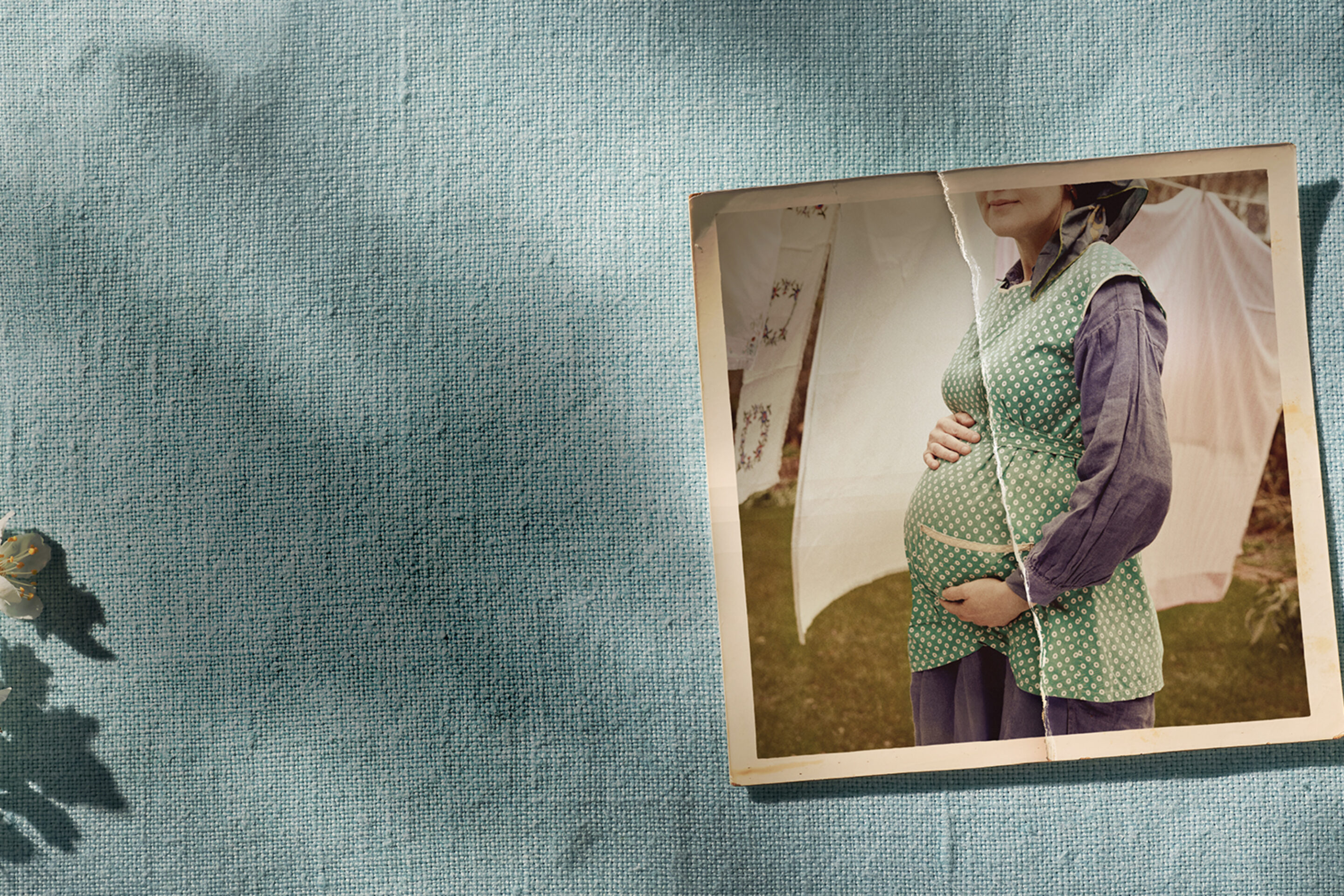 A vintage photo of a pregnant woman holding her bump is placed on a textured blue fabric background with shadows and white flowers in the corner.