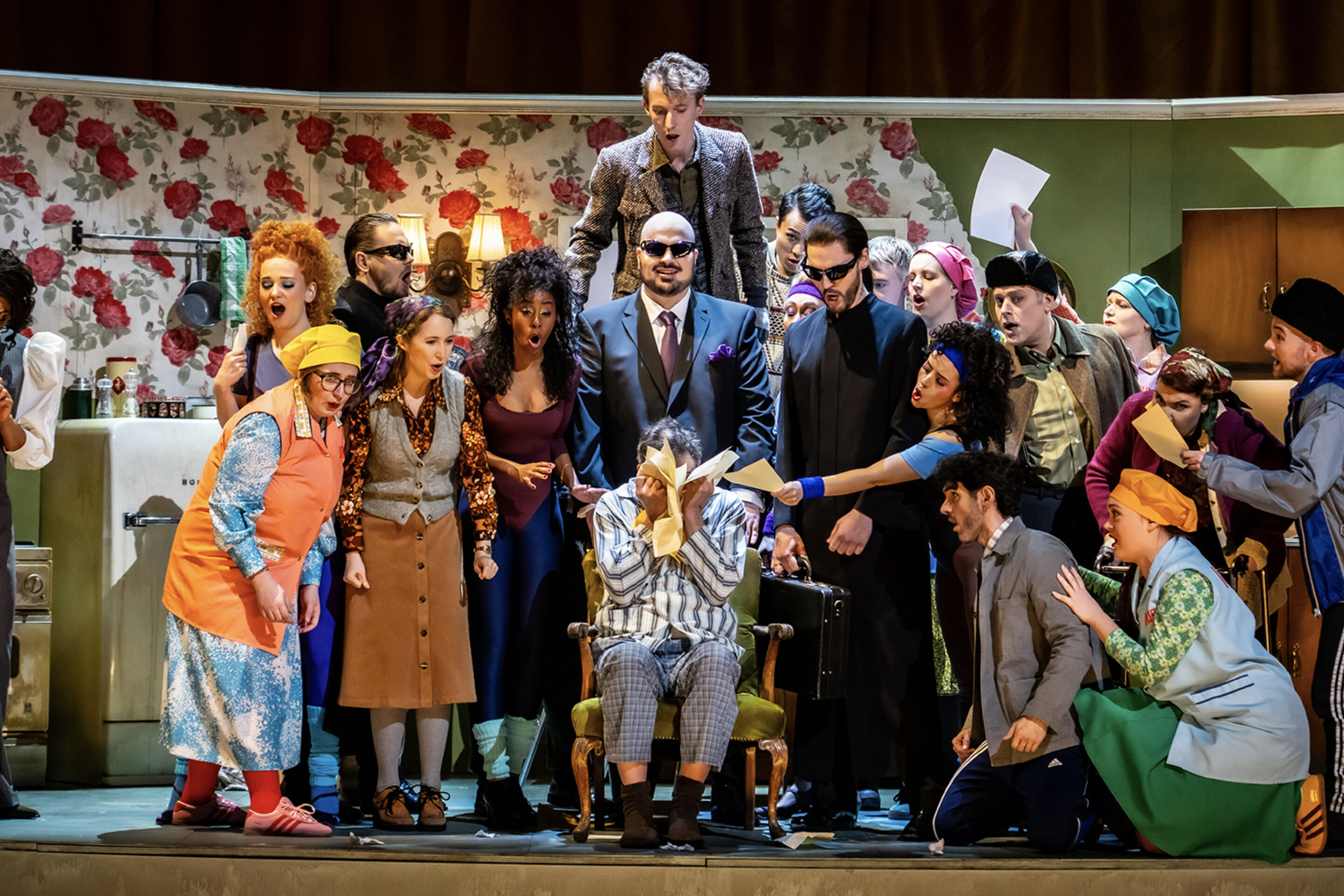 A lively comedy scene shows a group of actors in colourful costumes crowding around a person seated in a barber's chair, set in a retro kitchen with floral wallpaper. The group's animated expressions make the moment truly To Die For.