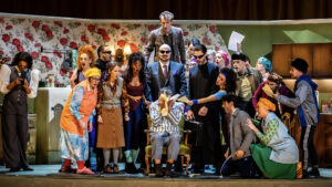 A lively comedy scene shows a group of actors in colourful costumes crowding around a person seated in a barber's chair, set in a retro kitchen with floral wallpaper. The group's animated expressions make the moment truly To Die For.