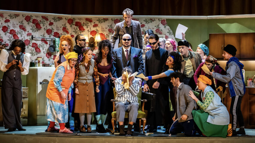 A lively comedy scene shows a group of actors in colourful costumes crowding around a person seated in a barber's chair, set in a retro kitchen with floral wallpaper. The group's animated expressions make the moment truly To Die For.