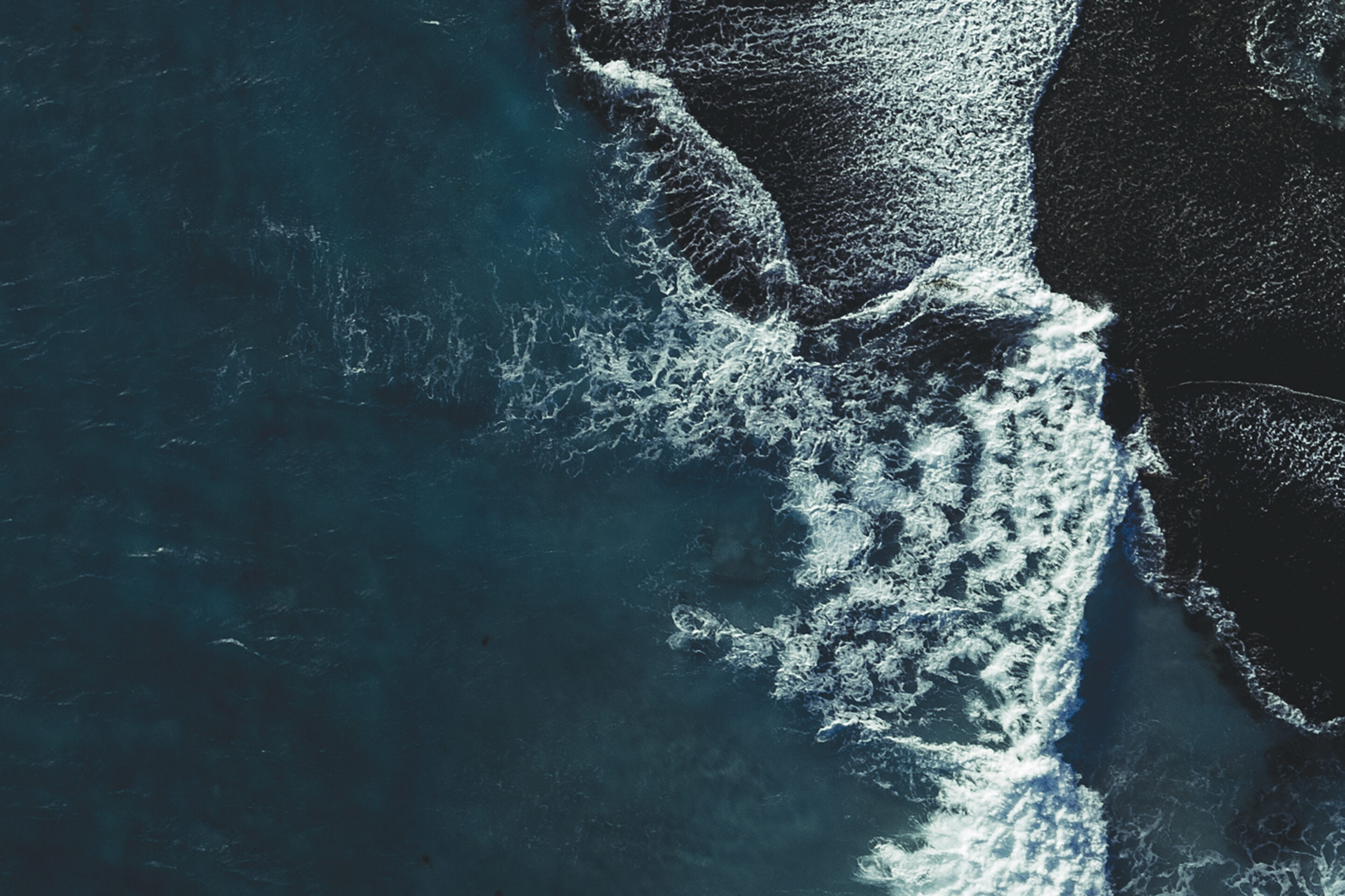 Aerial view of ocean waves crashing against dark, rocky shoreline, creating white foam and contrasting textures between deep blue water and rugged coast.