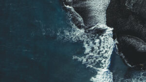 Aerial view of ocean waves crashing against dark, rocky shoreline, creating white foam and contrasting textures between deep blue water and rugged coast.