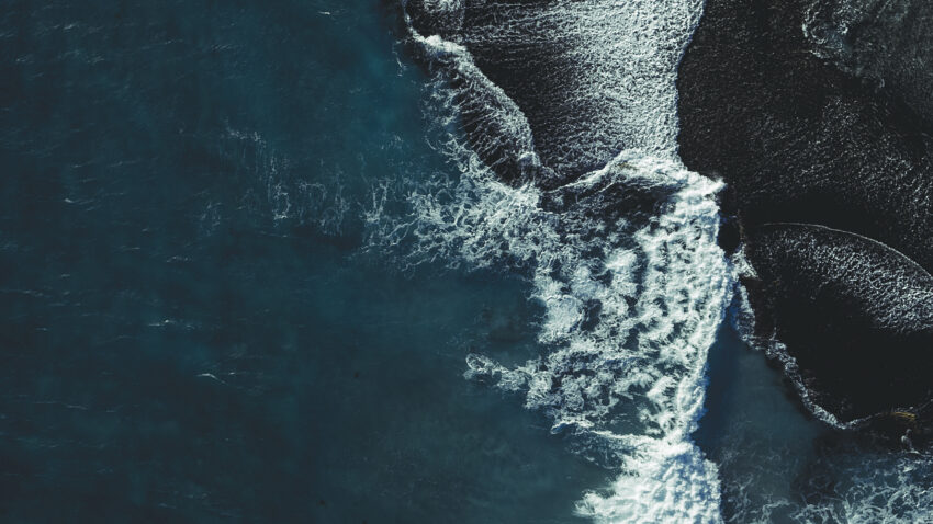 Aerial view of ocean waves crashing against dark, rocky shoreline, creating white foam and contrasting textures between deep blue water and rugged coast.