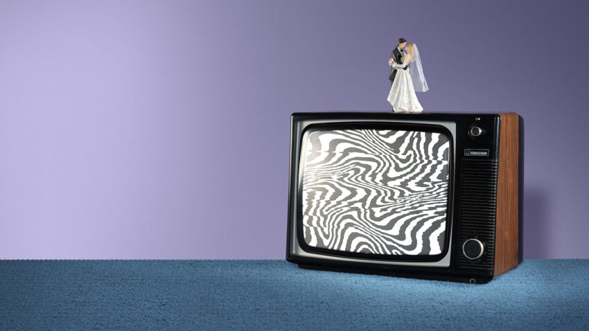 A bride and groom figurine stand on top of a vintage television displaying black and white static, against a purple wall and blue carpeted floor.