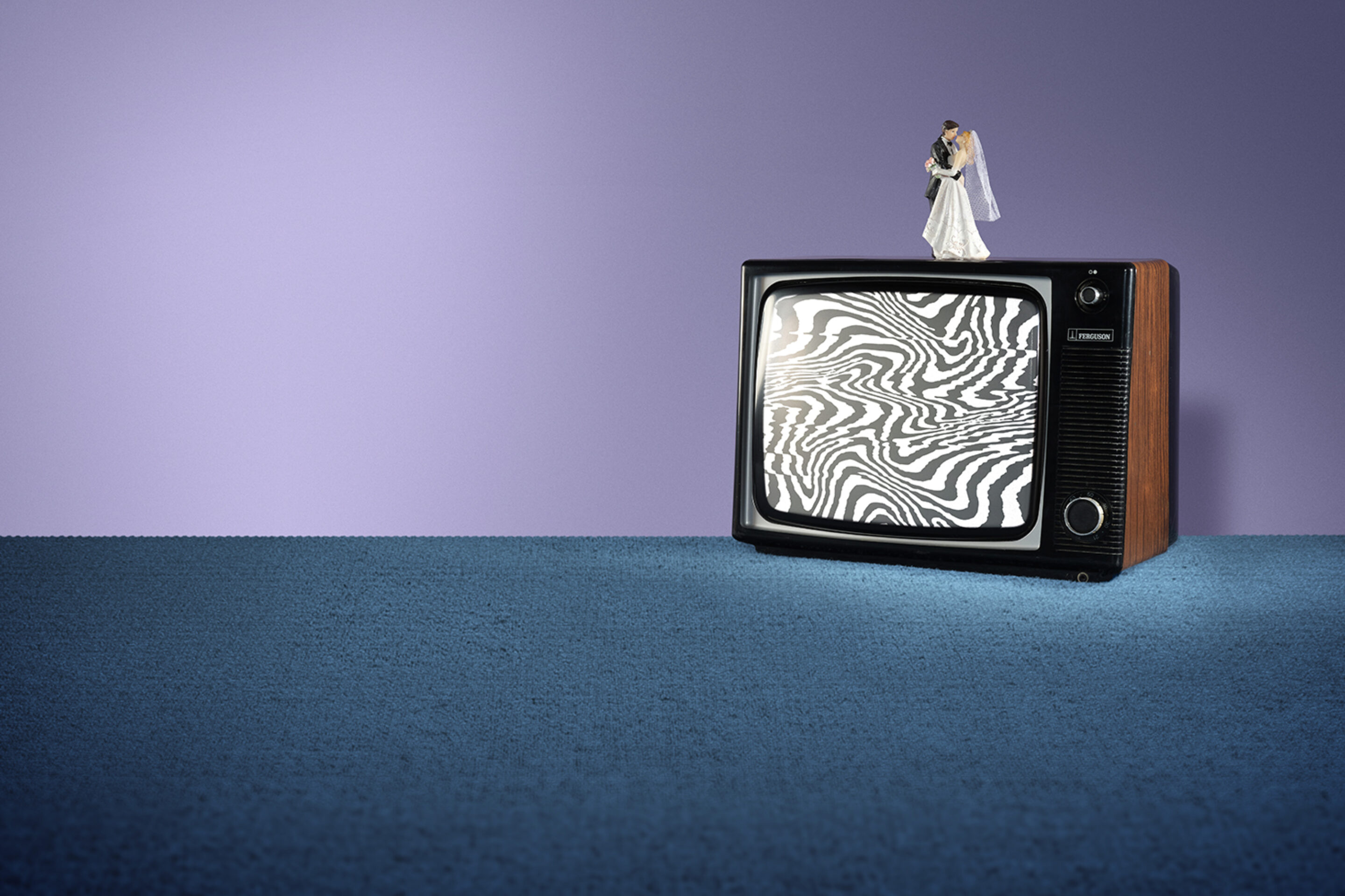 A vintage telly with a swirling black-and-white static screen sits on a blue carpet. On top of the telly, a small bride and groom figurine evoke scenes from an operetta. The background wall is purple.