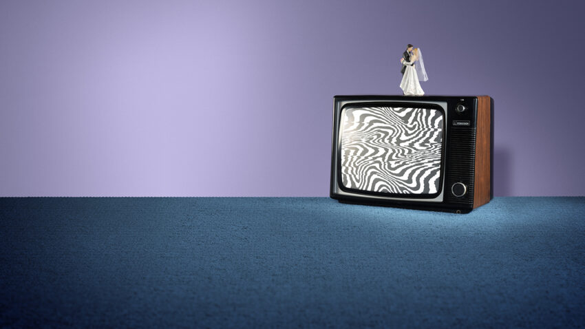 A vintage telly with a swirling black-and-white static screen sits on a blue carpet. On top of the telly, a small bride and groom figurine evoke scenes from an operetta. The background wall is purple.