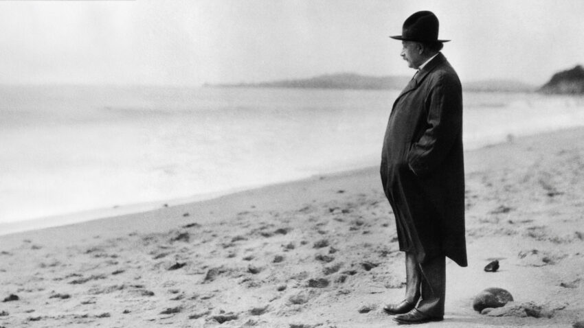 A man in a long coat and bowler hat stands alone on a sandy beach, looking out at the sea under a cloudy sky. The black-and-white scene evokes the contemplative mood of Philip Glass’s opera Einstein on the Beach.