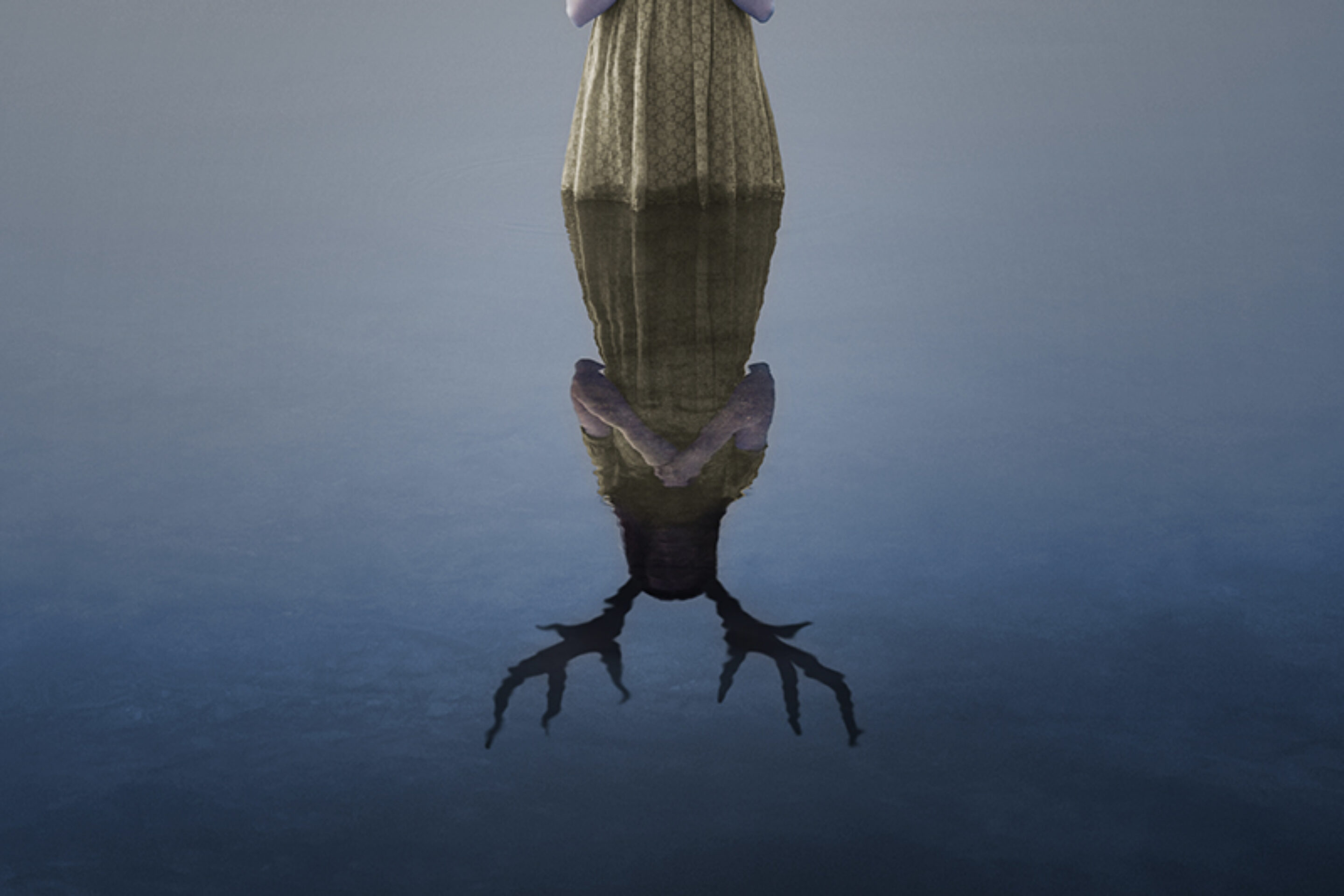 A person in a dress stands at the edge of calm water; their reflection reveals a stag’s antlers and head, evoking the surreal, dreamlike atmosphere of Iphigénie en Tauride.