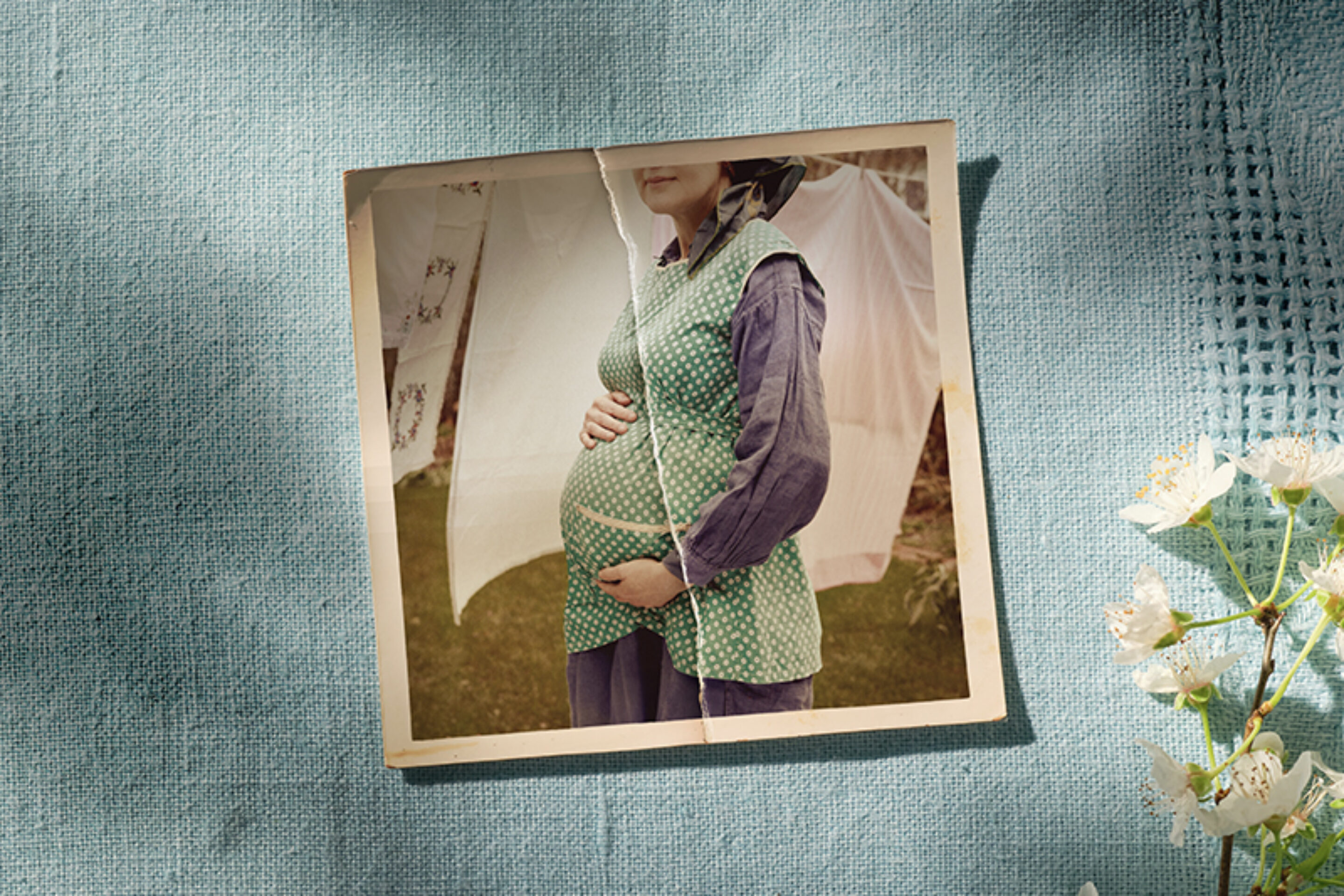 A vintage photo of a pregnant woman wearing a green polka dot waistcoat and blue dress, holding her belly, is placed on textured blue fabric next to blossoming white flowers. The woman's face is partially out of frame.