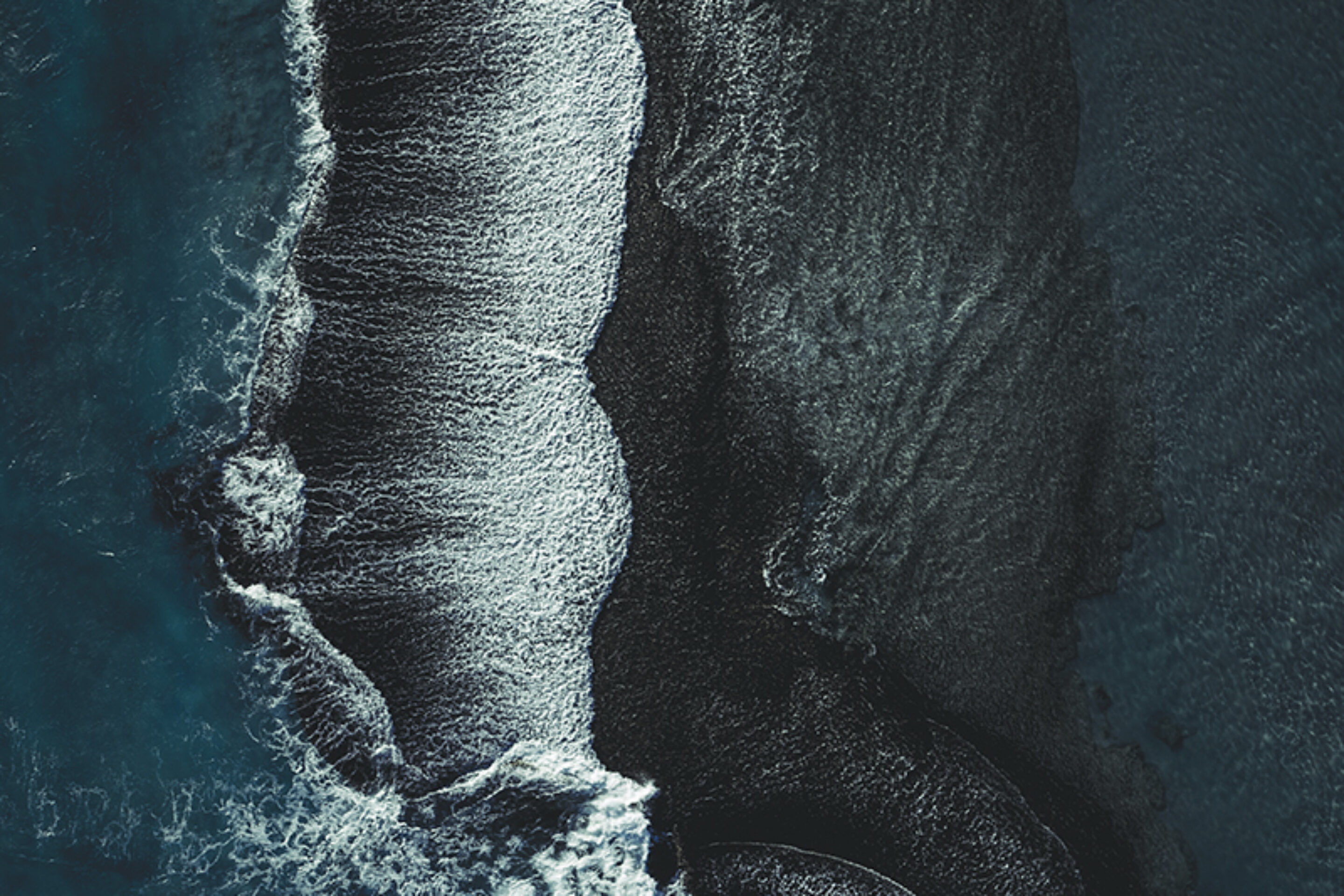 Aerial view of ocean waves crashing onto dark, textured sand, creating striking white foam patterns and deep contrasts along the shore.